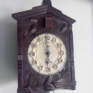 May include: A brown wooden cuckoo clock with a white face and black hands. The clock has a carved design of pine branches and cones. The clock is hanging from a chain.