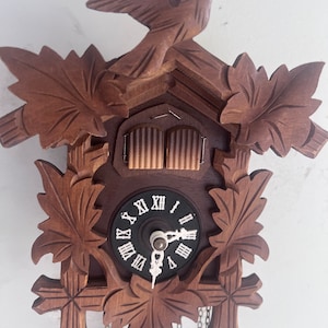 May include: A wooden cuckoo clock with a carved bird perched on top. The clock face is black with white Roman numerals. The clock is decorated with carved leaves and has hanging chains.