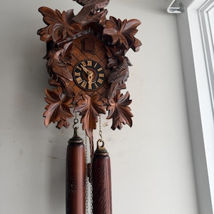 May include: A wooden cuckoo clock with carved birds and leaf accents. The clock face has Roman numerals and gold hands. Two dark brown pendulum weights hang below the clock, attached by chains. The clock is mounted on a light gray wall.