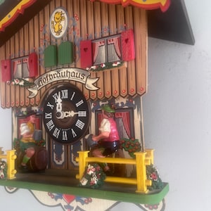 Può includere: Un orologio a cucù in legno con una facciata marrone e gialla. Il quadrante dell'orologio ha numeri romani e uno sfondo nero. L'orologio presenta due figure in abiti tradizionali tedeschi, che tengono boccali e barili. L'orologio ha un cartello con la scritta "Hofbräuhaus".