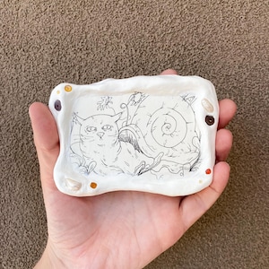 Puede incluir: Un cuenco de cerámica blanco con una ilustración dibujada a mano de un gato y un caracol. El cuenco tiene forma rectangular con bordes redondeados y está decorado con pequeños adornos de colores.
