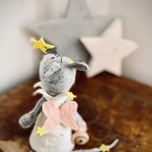 May include: A gray felt mouse with pink wings and yellow stars, looking up at a large gray star. The mouse is wearing a white dress and has a yellow star on its head.