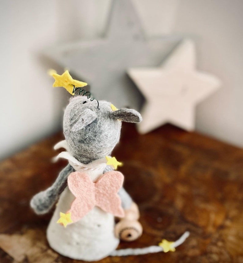 May include: A gray felt mouse with pink wings and yellow stars, looking up at a large gray star. The mouse is wearing a white dress and has a yellow star on its head.