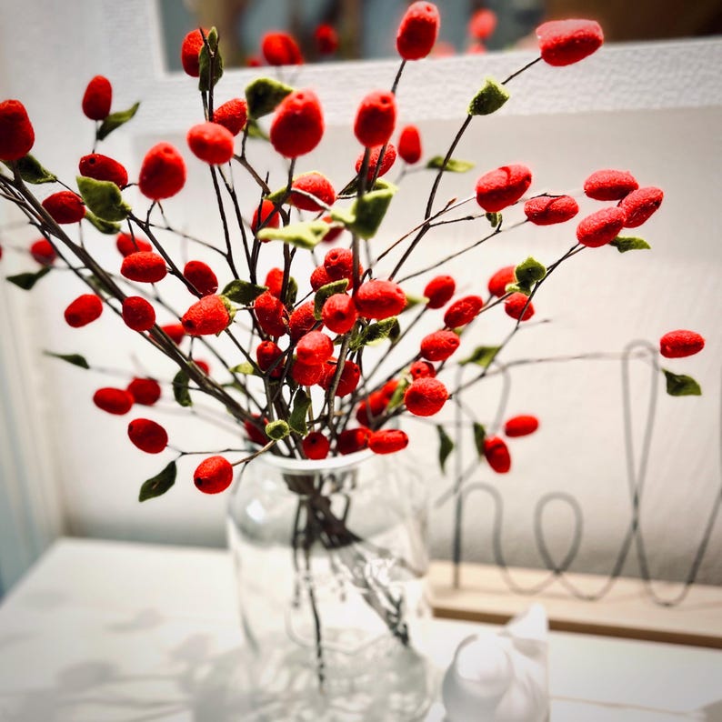 May include: A clear glass vase containing a decorative arrangement of thin, dark brown branches adorned with numerous small, round, bright red felt berries and tiny, irregular green felt leaves attached to the stems throughout the bouquet.