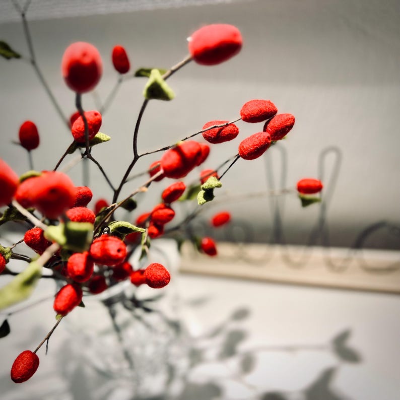 May include: A decorative branch featuring numerous small, vibrant red berries and tiny, textured green leaves. The branch is arranged with thin, dark brown stems that extend outward, creating a natural, organic appearance with bright color contrast.