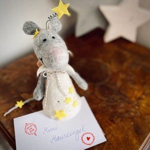 May include: A gray felt mouse with a white felt dress and yellow felt stars. The mouse is wearing a yellow felt star on its head. The mouse is sitting on a white card with the text "Mimi M&auml;useengel" written in black ink.