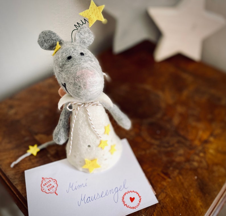 May include: A gray felt mouse with a white felt dress and yellow felt stars. The mouse is wearing a yellow felt star on its head. The mouse is sitting on a white card with the text "Mimi M&auml;useengel" written in black ink.