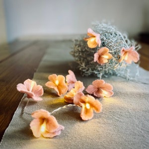 Könnte beinhalten: Eine Lichterkette mit rosa Filzblumen. Die Lichter sind mit einem dünnen Draht verbunden und leuchten. Die Blumen sind in einer Gruppe auf einer hellbraunen Oberfläche angeordnet.