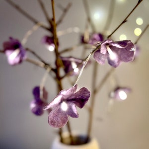 Könnte beinhalten: Eine dekorative Lichterkette mit kleinen, weichen, lavendelfarbenen Filzblumen, die an einem dünnen, flexiblen Draht befestigt sind. Die Blumenlichter sind mit kahlen, braunen Zweigen verflochten und bilden ein leuchtendes Arrangement aus violetten Blüten.
