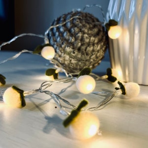 Könnte beinhalten: Eine LED-Lichterkette mit kleinen, runden, weißen Filzkugeln als Beeren und dunkelgrünen Filzblättern. Die Lichter strahlen ein warmes Licht aus und sind mit einem dünnen, flexiblen silbernen Draht verbunden, was für eine sanfte Beleuchtung sorgt.