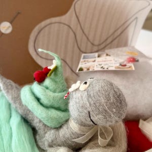 Könnte beinhalten: Eine nadelgefilzte graue Schneckenfigur mit einem mintgrünen Gehäuse, das mit roten Beeren verziert ist und ein weißes Band um den Hals trägt. Die Schnecke liegt in einer Bastelbox mit loser mintgrüner und grauer Wolle, einer Anleitung und rotem Filz.