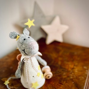 May include: A gray felt mouse with a white felt dress and yellow felt stars. The mouse is wearing a pink nose and has a white felt halo with a yellow felt star. The mouse is standing on a wooden surface.