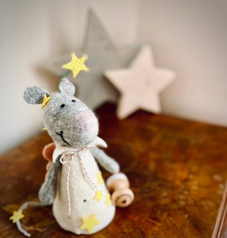 May include: A gray felt mouse with a white felt dress and yellow felt stars. The mouse is wearing a pink nose and has a white felt halo with a yellow felt star. The mouse is standing on a wooden surface.