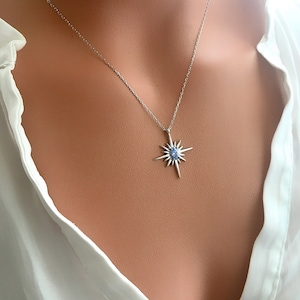 May include: A silver necklace with a starburst pendant featuring a light blue gemstone at its center. The pendant has multiple pointed rays extending outwards. The necklace is displayed against a white blouse.