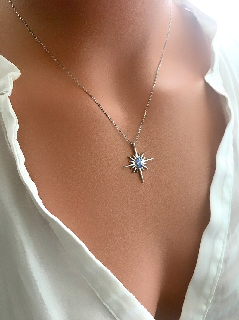 May include: A silver necklace with a starburst pendant featuring a light blue gemstone at its center. The pendant has multiple pointed rays extending outwards. The necklace is displayed against a white blouse.