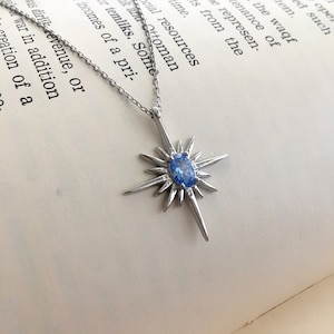 May include: A silver-toned necklace featuring a starburst pendant with a central, oval-shaped blue gemstone. The pendant has multiple pointed rays extending outwards. The necklace is displayed on a book with printed text.