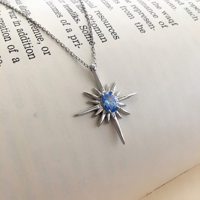 May include: A silver-toned necklace featuring a starburst pendant with a central, oval-shaped blue gemstone. The pendant has multiple pointed rays extending outwards. The necklace is displayed on a book with printed text.