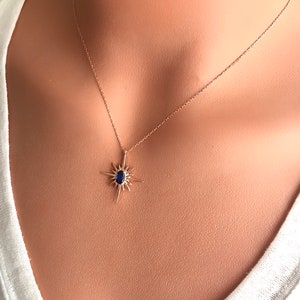 K&ouml;nnte beinhalten: Eine zarte goldene Halskette mit einem Sternburst-Anh&auml;nger, der einen blauen Edelstein in der Mitte tr&auml;gt.