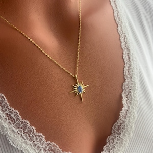 May include: A gold-colored necklace with a starburst pendant featuring a blue gemstone center. The necklace is worn against a white ribbed top with lace trim.