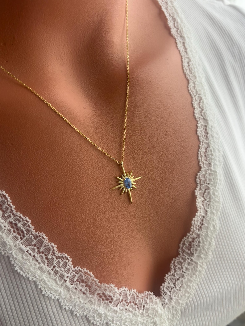 May include: A gold-colored necklace with a starburst pendant featuring a blue gemstone center. The necklace is worn against a white ribbed top with lace trim.
