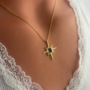 K&ouml;nnte beinhalten: Eine goldene Halskette mit einem Sternburst-Anh&auml;nger mit einem blauen Edelstein. Die Halskette tr&auml;gt ein Model, das ein wei&szlig;es Hemd mit Spitzenbesatz tr&auml;gt.