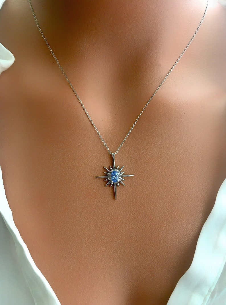 May include: A silver necklace featuring a starburst pendant with a central blue gemstone. The pendant has pointed rays extending outwards from a central point. The necklace is displayed against a skin tone background.