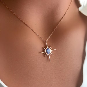May include: A delicate gold-colored necklace featuring a starburst pendant with a central blue gemstone. The pendant has multiple pointed rays extending outwards. The necklace is displayed on a person wearing a white shirt.