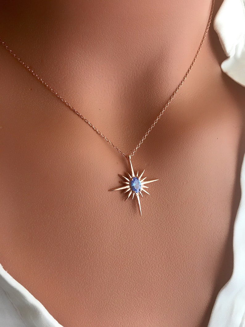 May include: A delicate gold-colored necklace featuring a starburst pendant with a central blue gemstone. The pendant has multiple pointed rays extending outwards. The necklace is displayed on a person wearing a white shirt.
