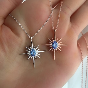 May include: Two necklaces with starburst pendants. One is silver-toned, the other is rose gold. Each pendant features a central, oval-shaped, light blue gemstone. The necklaces are displayed against a neutral background.