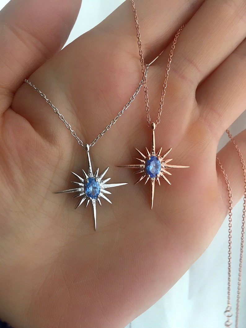 May include: Two necklaces with starburst pendants. One is silver-toned, the other is rose gold. Each pendant features a central, oval-shaped, light blue gemstone. The necklaces are displayed against a neutral background.
