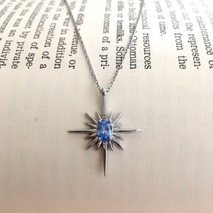 May include: A silver-toned necklace with a starburst pendant featuring a light blue oval gemstone at its center. The pendant has sharp, pointed rays extending outwards. The necklace rests on an open book.
