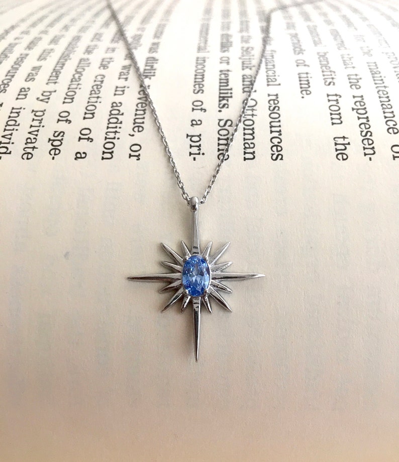 May include: A silver-toned necklace with a starburst pendant featuring a light blue oval gemstone at its center. The pendant has sharp, pointed rays extending outwards. The necklace rests on an open book.