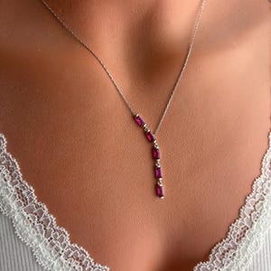 May include: A silver necklace featuring a series of five rectangular, deep pink gemstones. The gemstones are vertically aligned and suspended from a delicate chain. The necklace is displayed on a person wearing a white top with lace trim.