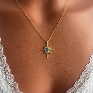 May include: A gold-colored necklace featuring a starburst pendant with a blue gemstone center. The necklace is displayed on a person wearing a white top with lace trim. The pendant has a sunburst design.
