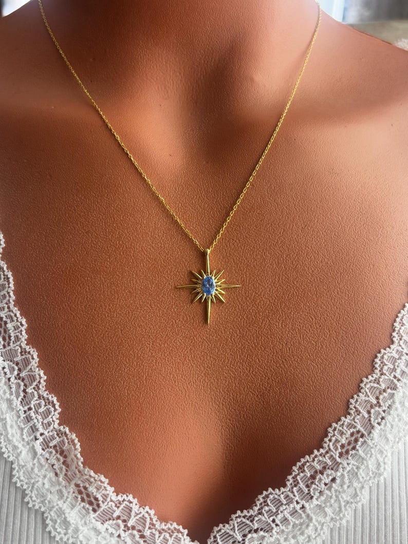May include: A gold-colored necklace featuring a starburst pendant with a blue gemstone center. The necklace is displayed on a person wearing a white top with lace trim. The pendant has a sunburst design.
