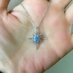 May include: A silver necklace featuring a starburst pendant with a central oval-cut, light blue gemstone. The pendant has pointed rays extending outwards. The necklace is displayed against a neutral background.