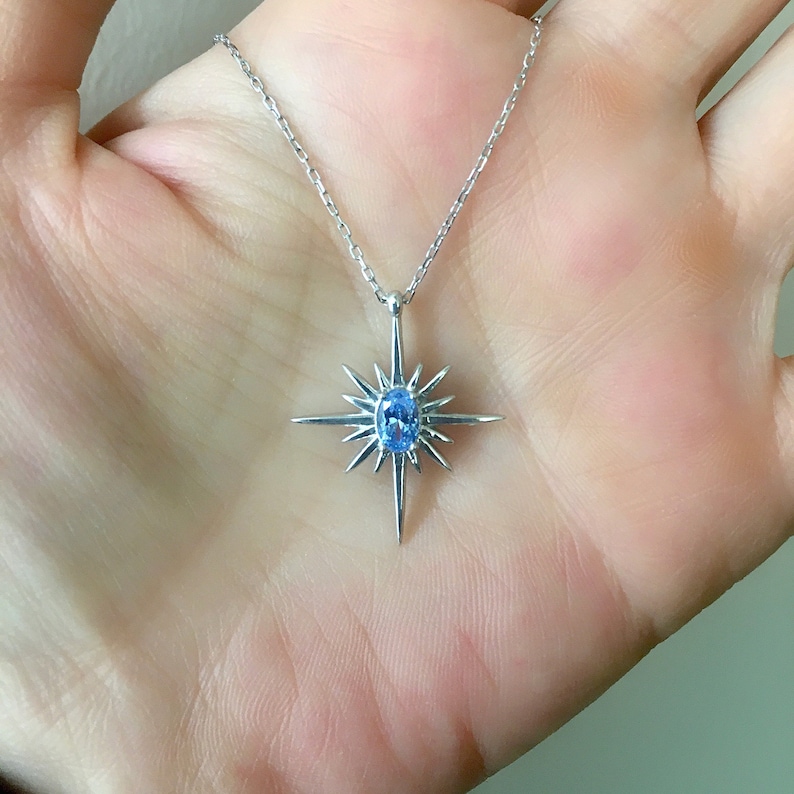 May include: A silver necklace featuring a starburst pendant with a central oval-cut, light blue gemstone. The pendant has pointed rays extending outwards. The necklace is displayed against a neutral background.