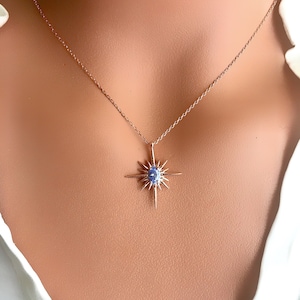 May include: A rose gold necklace featuring a starburst pendant with a central blue gemstone. The pendant has multiple pointed rays extending outwards. The necklace is displayed against a neutral background.
