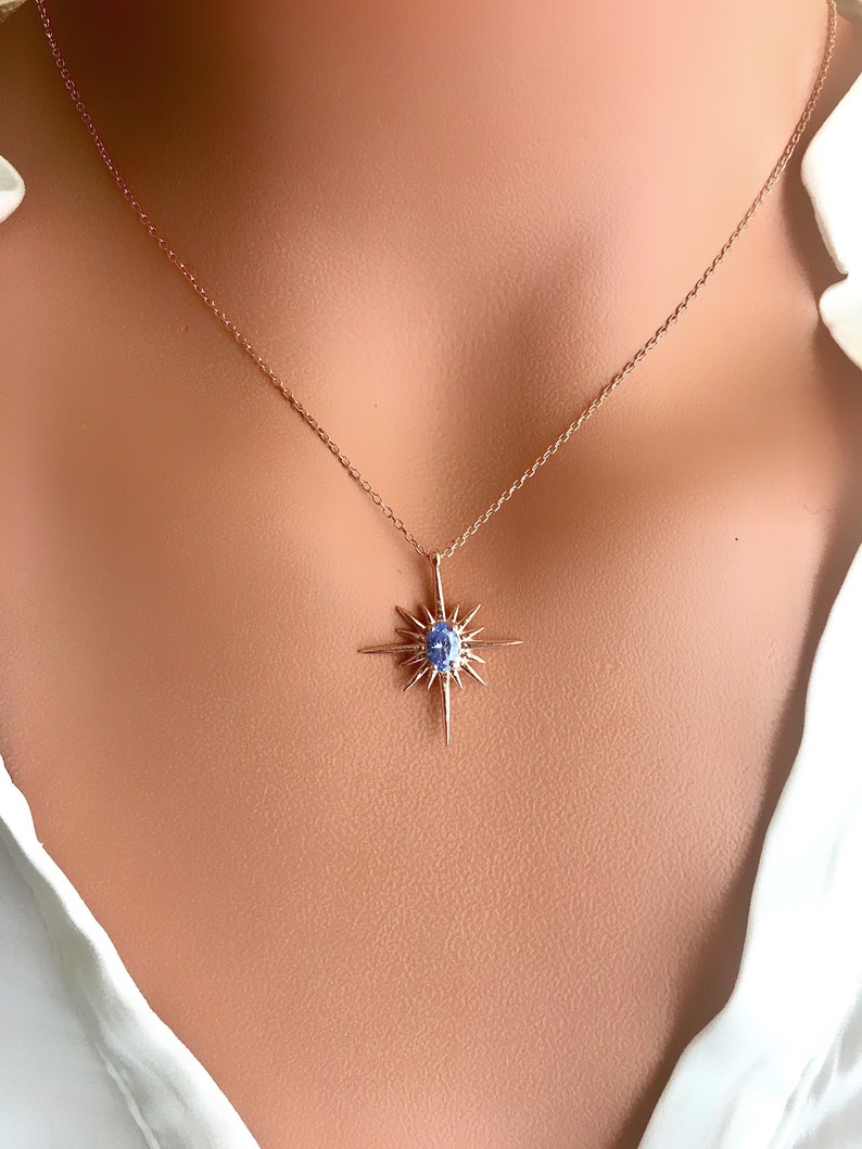 May include: A rose gold necklace featuring a starburst pendant with a central blue gemstone. The pendant has multiple pointed rays extending outwards. The necklace is displayed against a neutral background.