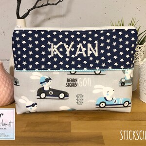 Puede incluir: Una bolsa con cremallera azul y blanca con un patr&oacute;n de estrellas y el nombre "KYAN" bordado en la parte delantera. La bolsa tiene un forro gris y blanco con un patr&oacute;n de conejos conduciendo coches de carreras. El texto "READY STEADY GO!" est&aacute; impreso en la bolsa.
