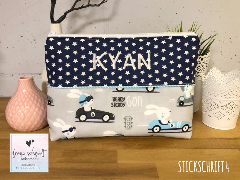 Puede incluir: Una bolsa con cremallera azul y blanca con un patr&oacute;n de estrellas y el nombre "KYAN" bordado en la parte delantera. La bolsa tiene un forro gris y blanco con un patr&oacute;n de conejos conduciendo coches de carreras. El texto "READY STEADY GO!" est&aacute; impreso en la bolsa.