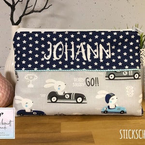 Puede incluir: Una bolsa con cremallera azul y blanca con una parte superior de tela azul marino con estrellas blancas y el nombre "JOHANN" bordado en blanco. La tela inferior es gris con un dise&ntilde;o de coche de carreras de conejo blanco y el texto "READY STEADY GO!!".