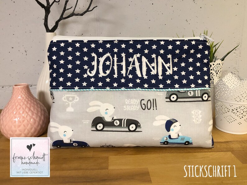 Puede incluir: Una bolsa con cremallera azul y blanca con una parte superior de tela azul marino con estrellas blancas y el nombre "JOHANN" bordado en blanco. La tela inferior es gris con un dise&ntilde;o de coche de carreras de conejo blanco y el texto "READY STEADY GO!!".