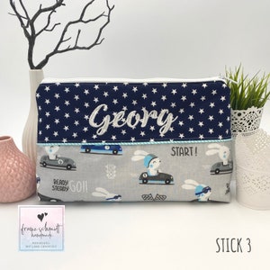 Puede incluir: Una bolsa con cremallera azul y blanca con una tela azul marino con estrellas blancas y el nombre "Georg" bordado en blanco. La bolsa tiene una tela gris con un patr&oacute;n de conejos conduciendo coches de carreras. El texto "START!" est&aacute; impreso en la bolsa. La bolsa est&aacute; hecha a mano y personalizada.