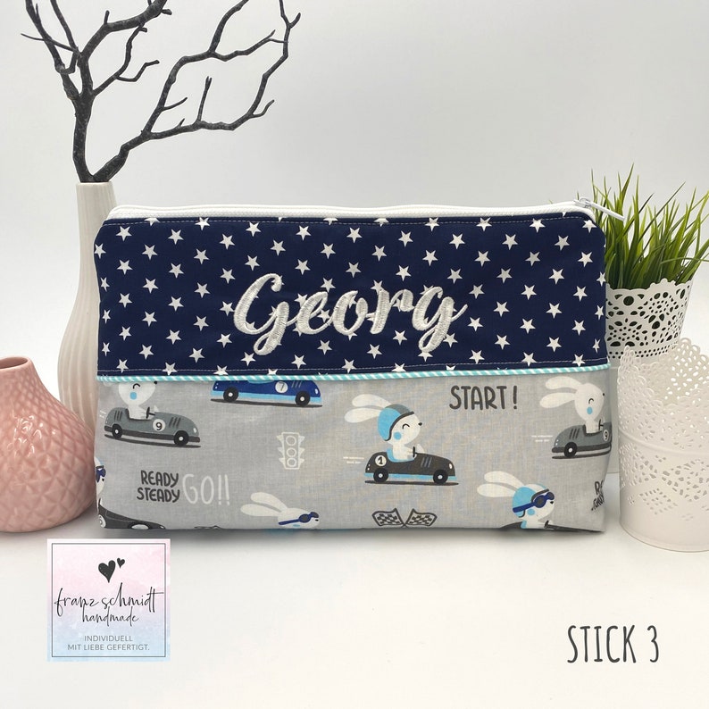 Puede incluir: Una bolsa con cremallera azul y blanca con una tela azul marino con estrellas blancas y el nombre "Georg" bordado en blanco. La bolsa tiene una tela gris con un patr&oacute;n de conejos conduciendo coches de carreras. El texto "START!" est&aacute; impreso en la bolsa. La bolsa est&aacute; hecha a mano y personalizada.