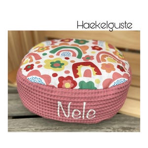 Könnte beinhalten: Ein rundes Bodenkissen mit einer weißen Oberseite, die mit einem bunten Muster aus Regenbögen, Blumen und Punkten bedruckt ist. Die Seiten bestehen aus altrosa Waffelstoff, auf dessen Vorderseite der Name Nele in weißem Garn gestickt ist.