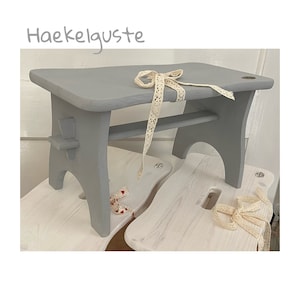 Könnte beinhalten: Ein grauer Holztritt mit rechteckiger Oberseite und gebogenen Beinen. Ein cremefarbenes Spitzenband ist über die Oberseite drapiert. Der Text "Haekelguste" befindet sich in der oberen linken Ecke. Der Hocker ist etwa 30 cm lang.