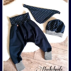 May include: A navy blue baby outfit set featuring a pair of harem pants, a bandana bib, and a beanie hat. The pants have striped cuffs, and the bib and hat are patterned with white anchors. The set is displayed on a white fluffy surface.