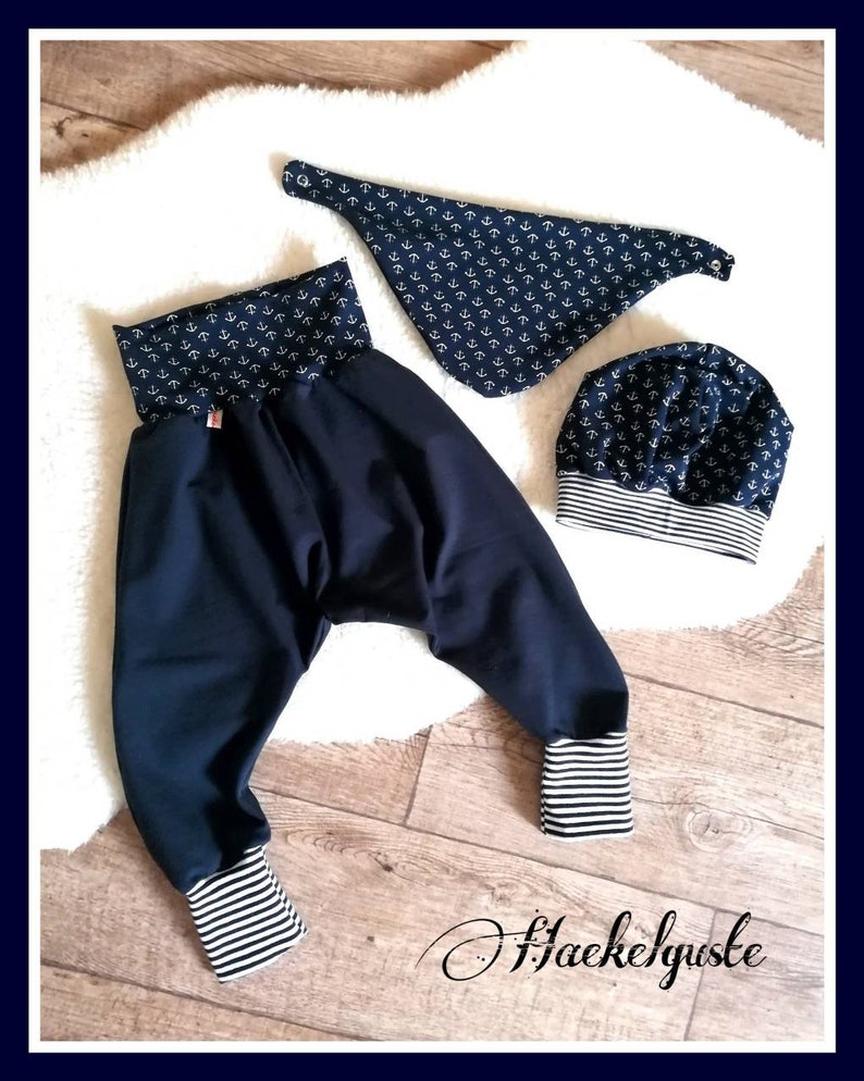 May include: A navy blue baby outfit set featuring a pair of harem pants, a bandana bib, and a beanie hat. The pants have striped cuffs, and the bib and hat are patterned with white anchors. The set is displayed on a white fluffy surface.