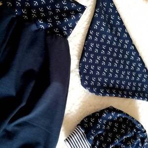 May include: Navy blue fabric items with a white anchor pattern. The items include a pair of pants, a bib, and a beanie. The beanie has a white and navy blue striped cuff. The items are on a white, fluffy surface.